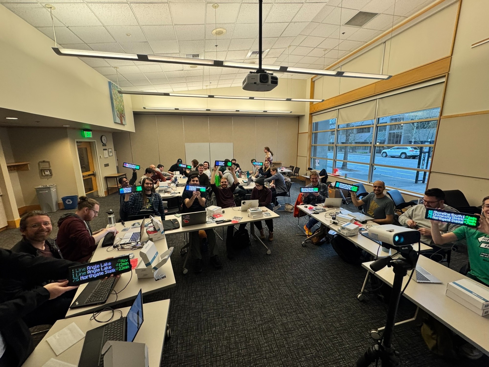 A group of people in a conference room at Redmond Library, sitting at tables with laptops and electronic components. Many are smiling and holding up Transit Trackers displaying arrival times for bus routes. A projector is set up in the foreground, and large windows show a street with parked cars outside.